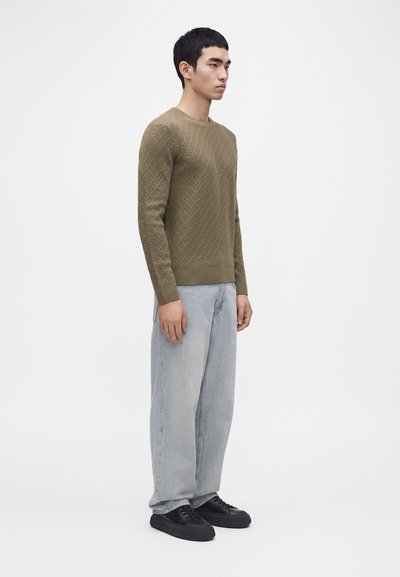 Homme debout de côté portant un pull texturé vert olive, un jean bleu clair ample et des baskets noires sur fond blanc uni.