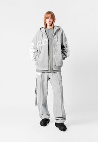 Sudadera gris con acentos de logo blancos, combinada con pantalones de chándal gris a juego. El conjunto está hecho de una tela suave con un corte relajado. Zapatos negros texturizados.