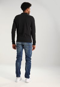 Zwarte longsleeve shirt gecombineerd met blauwe jeans. Het shirt heeft een eenvoudig ontwerp, terwijl de jeans een vervaagde textuur hebben met details op de achterzak.