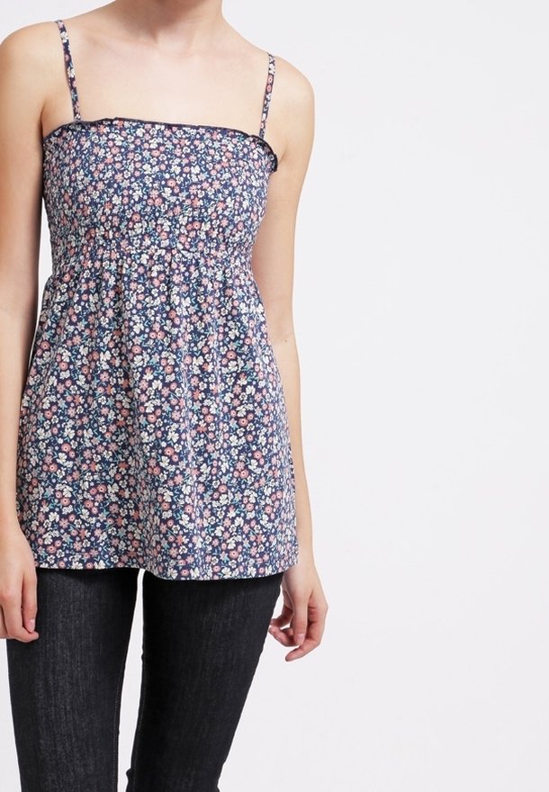 Femme portant un haut bleu fleuri sans manches avec de fines bretelles et un pantalon noir sur un fond clair uni.