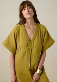 Robe jaune moutarde en tissu texturé, décolleté en V, manches courtes et taille à cordon. Accentuée d'une bordure décorative et de pompons.