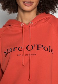 Felpa rossa con cappuccio regolabile, polsini a coste e stampa grafica frontale con il testo "Marc O'Polo EST. A STOCCOLMA."