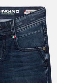 Tmavě modré džínové kalhoty s viditelným šitím a malým italským praporem na kapse. Obsahují kovový knoflík na pase.