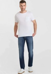 T-shirt blanc en coton avec une poche poitrine, associé à un jean en denim bleu foncé et des baskets blanches avec des semelles en caoutchouc marron.