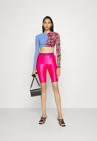 Barevný cropped top s dlouhým rukávem, zdobený modrými a růžovými vzory, doplněný lesklými růžovými biker šortkami. Ozdoben černou kabelkou a sandály.