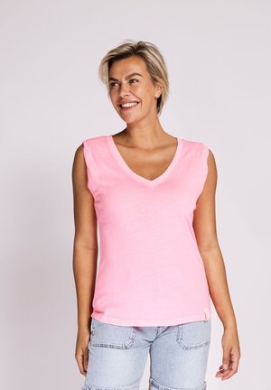 Lächelnde Frau mit kurzen blonden Haaren, die ein pinkfarbenes ärmelloses Top mit V-Ausschnitt und hellblaue Jeansshorts trägt und vor einem einfarbigen Hintergrund steht.