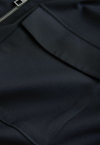 Schwarzer Stoff mit einer glatten, matten Textur, einem Reißverschlussdetail und einer gefalteten Tasche. Nähte sind sichtbar.