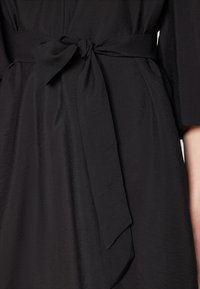 Svart klänning med texturerad yta, som har ett knytbart midjebälte och korta ärmar, vilket betonar tygets släthet och den minimalistiska designen.