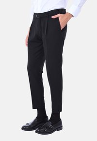 Pantaloni neri su misura con pieghe, chiusura frontale con bottone e tasche laterali. Abbinati a mocassini neri e calzini neri, completati da una camicia bianca.