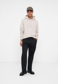 Felpa beige con cappuccio e tasca frontale, abbinata a pantaloni neri e scarpe nere. Accentuata da un cappello a scacchi marrone.