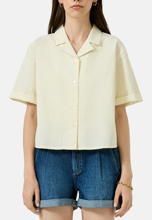 Frau trägt ein kurzärmeliges, blassgelbes Hemd mit einem dezenten Karomuster und hochgekrempelte blaue Jeansshorts.