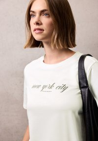T-shirt en coton blanc avec le texte vert "new york city" en lettres élégantes. Accentuée par un sac à épaule en cuir noir.
