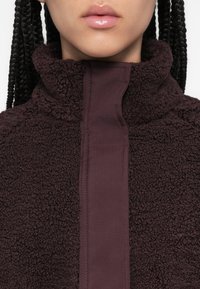 Maronne Fleecejacke mit hohem Kragen, strukturierter Oberfläche und einem kontrastierenden, glatten Einsatz an der Vorderseite. Verfügt über ein Reißverschlussdetail.