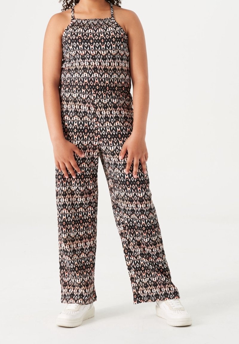 Dziewczyna ubrana w czarno-biało-różowy kombinezon bez rękawów i białe trampki, stoi na tle jednolitego tła.