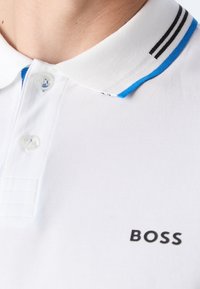 Polo blanc avec une patte de boutonnage à deux boutons, un col rayé noir et bleu, et un logo noir sur le côté gauche. Tissu texturé.