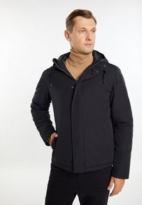 Sort isoleret jakke med hætte, med lynlås foran, sidelommer og en glat tekstur. Båret over en lys brun turtleneck.