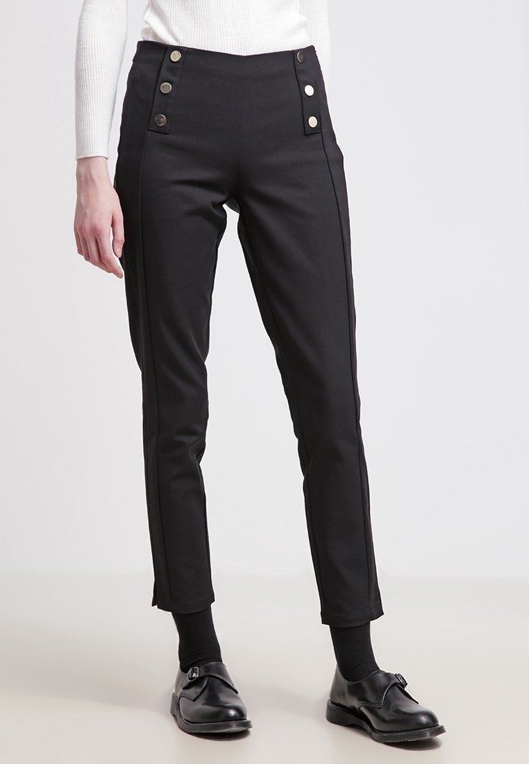 Femme portant un pantalon noir slim avec des détails à boutons près de la taille, des chaussettes noires et des chaussures noires à boucle, debout devant un fond uni.