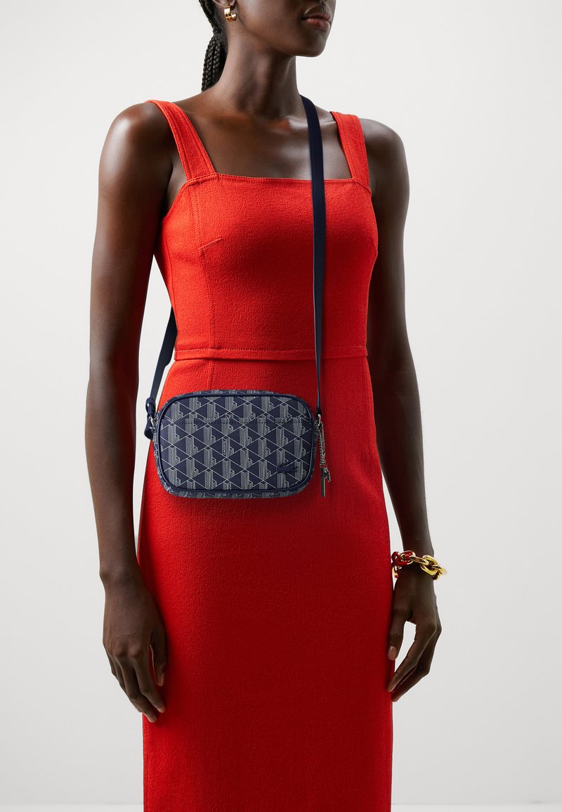 Borsa a tracolla blu navy con motivo geometrico, chiusura a zip e tracolla regolabile, indossata sopra un vestito rosso a trama.