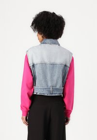 Giacca di jeans con un lavaggio azzurro chiaro, design cropped e una texture di denim, abbinata a un top a maniche lunghe di un rosa brillante sotto.