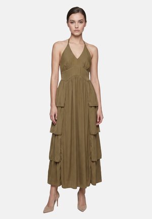 Donna che indossa un abito lungo verde oliva con collo halter e dettagli a volant sovrapposti, in piedi con espressione neutra e tacchi alti beige.