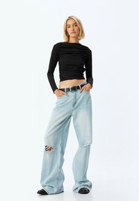 Sort langærmet top med samlet tekstur, parret med lyseblå bredbenede jeans med slidser og et sort bælte med en sølvspænde.