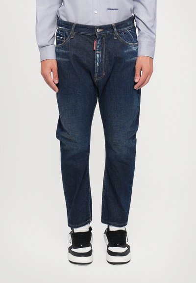 Jean en denim bleu foncé avec une coupe décontractée, décoloration visible et cinq poches. Associé à des baskets noires et blanches avec un tissu texturé.