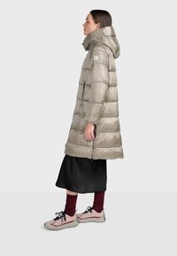 Lång silverfärgad vadderad jacka med hög krage, dragkedjefickor och glänsande yta. Bärs över en svart kjol och rosa sneakers med mörka strumpor.