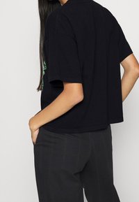 Černé kratké tričko z bavlny s kulatým výstřihem a krátkými rukávy. Obsahuje boční rozparky a volný střih. Zdobeno grafickým designem.