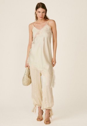 Mujer con vestido lencero color crema con encaje en el dobladillo sobre pantalones anchos con textura, sosteniendo un bolso tejido y usando sandalias de tacón, posando frente a un fondo neutro.