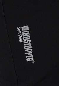 Schwarzer Stoff mit weißem, diagonal bedrucktem "WINDSTOPPER Soft Shell"-Branding, das den Materialtyp und die Marke bei Outerwear hervorhebt.