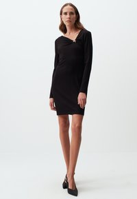 Zwarte jurk met lange mouwen en een aansluitende silhoutte, voorzien van een gedraaide halslijn en kleine metallic accenten, gemaakt van een geribbelde stof.