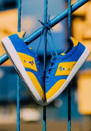 Blauwe en gele sneakers van suede en rubber. Heeft een gele streep, gestructureerde veters en witte rubberen zolen.