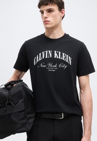 Νέος άνδρας που φοράει μαύρο T-shirt Calvin Klein με το κείμενο "New York City", κρατώντας μια μαύρη τσάντα duffel, μπροστά σε απλό ανοιχτό φόντο.