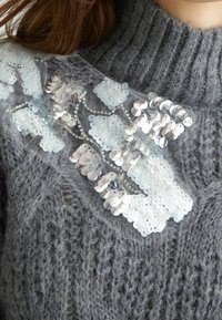 Grau gestrickter Pullover mit strukturiertem Muster. Hat ein transparentes Overlay, das mit silbernen und weißen Pailletten geschmückt ist, die in einem wellenförmigen Design angeordnet sind.