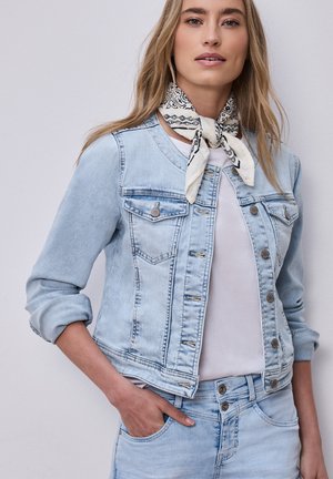 Femme portant une veste en jean bleu clair et un jean, une chemise blanche et un foulard blanc à motifs autour du cou, une main dans la poche.
