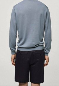 Mann in hellblauem Langarmpullover und schwarzen knielangen Shorts, mit dem Rücken zur Kamera vor einem einfarbigen Hintergrund stehend.