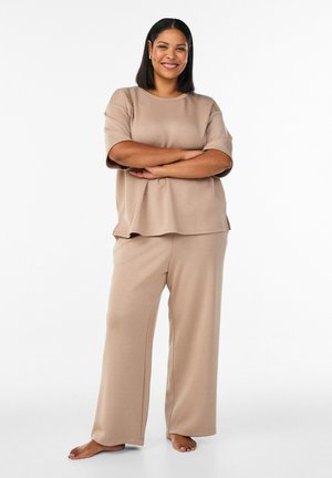 Sieviete stāv basām kājām ar sakrustotām rokām, ģērbta vaļīgā, īsrokainā bēšā topā un atbilstošās vaļējās biksēs ar plašām kājām uz vienkārša balta fona.