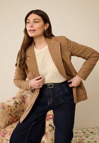 Bruine corduroy blazer met geborduurde accenten, gecombineerd met een crèmekleurige gebreide trui, high-waisted donker denim en een zwarte riem. Achtergrond met een bloemen fauteuil.