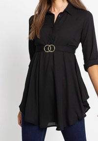 Blouse tunique noire avec un devant boutonné, des manches longues, une taille ceinturée avec un accent en double anneau doré et un ourlet léger et superposé.