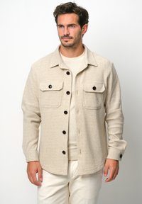 Giacca beige texture con maniche lunghe, due tasche sul petto e bottoni scuri. Indossata sopra un top beige chiaro, su uno sfondo bianco.