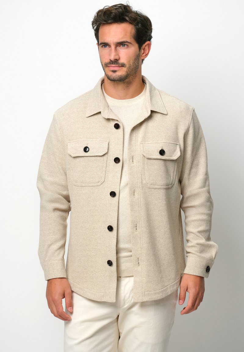 Giacca beige texture con maniche lunghe, due tasche sul petto e bottoni scuri. Indossata sopra un top beige chiaro, su uno sfondo bianco.