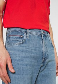 Vaqueros de mezclilla azul claro con una textura ligeramente descolorida, que cuentan con cinco bolsillos y acentos de costura roja en contraste. Llevados con una camisa roja.