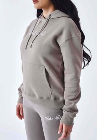 Lichtgrijze hoodie met een voorzak, verstelbare trekkoord en logodetails. Gecombineerd met bijpassende leggings met vergelijkbare branding.