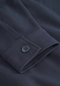 Tela texturizada de color azul oscuro con un patrón de cuadrícula, con un botón de plástico negro en el puño. El diseño general es funcional y contemporáneo.