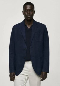 Marinblå kavaj med två knappar, slagkrage och bröstficka. Buren över en mörk stickad tröja, kombinerad med ljusa byxor.