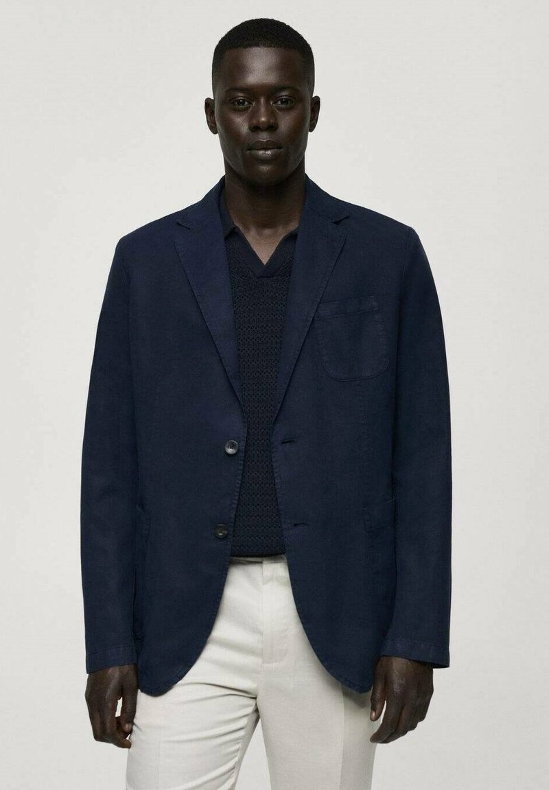 Marinblå kavaj med två knappar, slagkrage och bröstficka. Buren över en mörk stickad tröja, kombinerad med ljusa byxor.