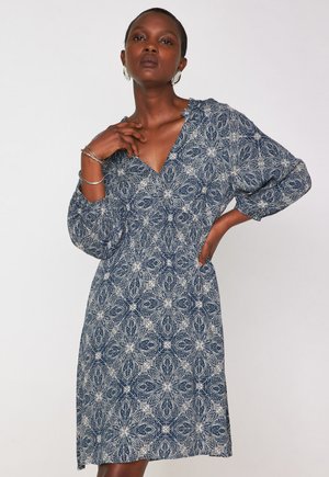 Donna che indossa un vestito midi blu con motivi floreali bianchi, maniche a tre quarti, e orecchini a cerchio e braccialetti in argento su uno sfondo semplice.