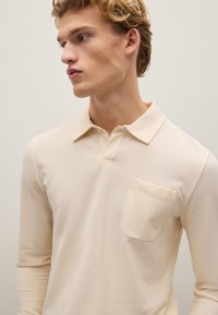 Langarm-Polohemd aus hellem beigen Baumwollstoff mit feiner Textur, ausgestattet mit einem klassischen Kragen und einer kleinen Brusttasche.