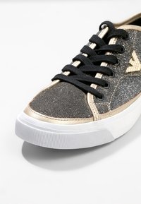 Czarny błyszczący materiałowy sneaker ze złotym wykończeniem, czarnymi sznurowadłami, białą gumową podeszwą i złotym logo z boku.