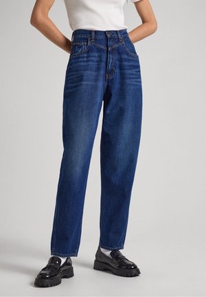 Person trägt hoch taillierte blaue Jeans, weißes Hemd und schwarze klobige Loafer, steht mit den Händen in den Taschen vor einem schlichten Hintergrund.